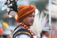 A person wearing traditional attire featuring a colorful turban with orange and red patterns, adorned with feathers. The individual is surrounded by blurred outdoor lighting. They hold a decorative object, possibly a ceremonial sword.