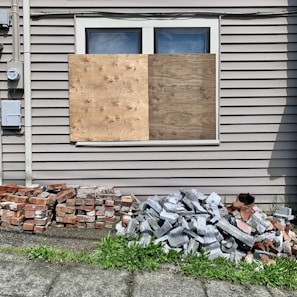 A building wall with two large wooden boards covering a window. Below the window, there are piles of bricks and concrete blocks. The siding on the building is beige, and there is a patch of green grass in the foreground.