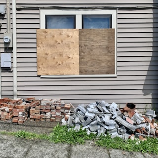 A building wall with two large wooden boards covering a window. Below the window, there are piles of bricks and concrete blocks. The siding on the building is beige, and there is a patch of green grass in the foreground.