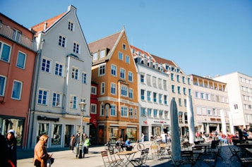 A lively urban square with colorful historic buildings in a European style lining the street. People are casually walking or sitting at outdoor cafes, enjoying a sunny day. The buildings are painted in shades of orange, blue, and beige, and feature traditional windows and rooftops. The area seems vibrant with a mix of locals and tourists.