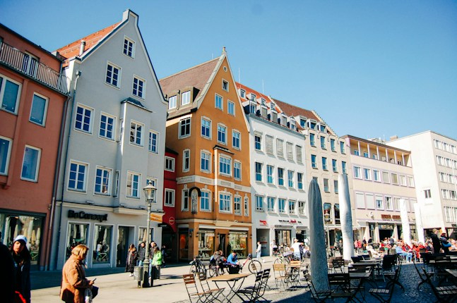 A lively urban square with colorful historic buildings in a European style lining the street. People are casually walking or sitting at outdoor cafes, enjoying a sunny day. The buildings are painted in shades of orange, blue, and beige, and feature traditional windows and rooftops. The area seems vibrant with a mix of locals and tourists.