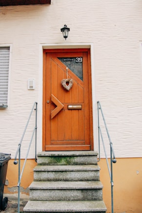 A wooden door with a geometric design, adorned with a heart-shaped wreath. It is placed at the top of a short staircase with three stone steps. A black lantern is mounted above the door, and a metal railing flanks the steps.