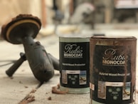 A close-up view of two cans of wood protector labeled 'Rubio Monocoat Hybrid Wood Protector' positioned on a concrete surface. Nearby, there is a power tool with a circular sanding attachment, suggesting the setting is related to woodworking or home improvement. The scene contains some wood shavings scattered around.