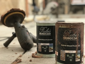 A close-up view of two cans of wood protector labeled 'Rubio Monocoat Hybrid Wood Protector' positioned on a concrete surface. Nearby, there is a power tool with a circular sanding attachment, suggesting the setting is related to woodworking or home improvement. The scene contains some wood shavings scattered around.