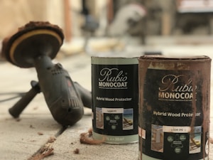 A close-up view of two cans of wood protector labeled 'Rubio Monocoat Hybrid Wood Protector' positioned on a concrete surface. Nearby, there is a power tool with a circular sanding attachment, suggesting the setting is related to woodworking or home improvement. The scene contains some wood shavings scattered around.
