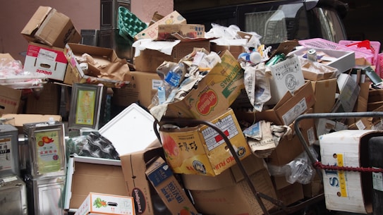 A large pile of cardboard boxes and packaging materials, including some metal containers and plastic items, appears to be discarded in a narrow space. Some of the boxes have visible brand labels and there are various shapes and sizes of packaging.