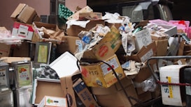 A large pile of cardboard boxes and packaging materials, including some metal containers and plastic items, appears to be discarded in a narrow space. Some of the boxes have visible brand labels and there are various shapes and sizes of packaging.