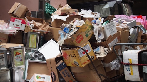 A large pile of cardboard boxes and packaging materials, including some metal containers and plastic items, appears to be discarded in a narrow space. Some of the boxes have visible brand labels and there are various shapes and sizes of packaging.