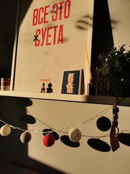 A shelf with various decorative items is lit by warm sunlight. On the shelf, a large piece of wall art with red lettering is prominently displayed. Below it, there is a smaller book or journal with the phrase 'Be Yourself'. Decorative string lights with ball-shaped covers hang in front of the shelf. A small plant and incense sticks are also arranged neatly on the shelf, adding to the cozy atmosphere.