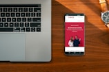 A wooden desk hosts a laptop on the left and a smartphone on the right, which displays an advertisement for a bank with the slogan 'Spend and save smartly'. A wristwatch lies above the phone, its metallic face and band accented by the warm tones of the wood.