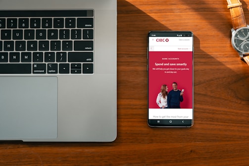 A wooden desk hosts a laptop on the left and a smartphone on the right, which displays an advertisement for a bank with the slogan 'Spend and save smartly'. A wristwatch lies above the phone, its metallic face and band accented by the warm tones of the wood.