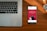 A wooden desk hosts a laptop on the left and a smartphone on the right, which displays an advertisement for a bank with the slogan 'Spend and save smartly'. A wristwatch lies above the phone, its metallic face and band accented by the warm tones of the wood.