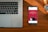 A wooden desk hosts a laptop on the left and a smartphone on the right, which displays an advertisement for a bank with the slogan 'Spend and save smartly'. A wristwatch lies above the phone, its metallic face and band accented by the warm tones of the wood.