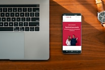 A wooden desk hosts a laptop on the left and a smartphone on the right, which displays an advertisement for a bank with the slogan 'Spend and save smartly'. A wristwatch lies above the phone, its metallic face and band accented by the warm tones of the wood.