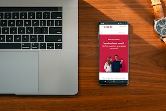 A wooden desk hosts a laptop on the left and a smartphone on the right, which displays an advertisement for a bank with the slogan 'Spend and save smartly'. A wristwatch lies above the phone, its metallic face and band accented by the warm tones of the wood.