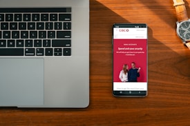 A wooden desk hosts a laptop on the left and a smartphone on the right, which displays an advertisement for a bank with the slogan 'Spend and save smartly'. A wristwatch lies above the phone, its metallic face and band accented by the warm tones of the wood.
