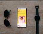 A smartphone displaying a promotional message is placed on a wooden surface. The screen shows a cheerful person with their hands behind their head, wearing a pink hat and sweatshirt against a vibrant yellow background. Text highlights a zero-fee feature and exclusive scholarship access. Beside the phone are a pair of black sunglasses and a black wristwatch, creating a balanced composition.