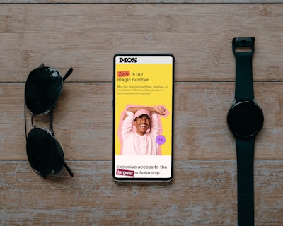 A smartphone displaying a promotional message is placed on a wooden surface. The screen shows a cheerful person with their hands behind their head, wearing a pink hat and sweatshirt against a vibrant yellow background. Text highlights a zero-fee feature and exclusive scholarship access. Beside the phone are a pair of black sunglasses and a black wristwatch, creating a balanced composition.