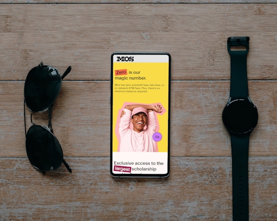 A smartphone displaying a promotional message is placed on a wooden surface. The screen shows a cheerful person with their hands behind their head, wearing a pink hat and sweatshirt against a vibrant yellow background. Text highlights a zero-fee feature and exclusive scholarship access. Beside the phone are a pair of black sunglasses and a black wristwatch, creating a balanced composition.