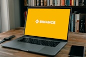 A laptop is positioned on a wooden table with an empty yellow screen displaying the word 'Binance' in white. Next to the laptop, a smartwatch and a smartphone are placed. In the background, there is a shelf filled with various books.