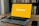 A laptop is positioned on a wooden table with an empty yellow screen displaying the word 'Binance' in white. Next to the laptop, a smartwatch and a smartphone are placed. In the background, there is a shelf filled with various books.