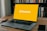 A laptop is positioned on a wooden table with an empty yellow screen displaying the word 'Binance' in white. Next to the laptop, a smartwatch and a smartphone are placed. In the background, there is a shelf filled with various books.