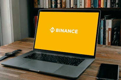 A laptop is positioned on a wooden table with an empty yellow screen displaying the word 'Binance' in white. Next to the laptop, a smartwatch and a smartphone are placed. In the background, there is a shelf filled with various books.