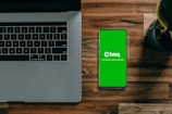 A laptop with a partially visible keyboard situated on a wooden surface next to a smartphone displaying a green screen with the word 'bisq' and the text 'Exchange, Decentralized'. There is also a small potted plant in the top-right corner.