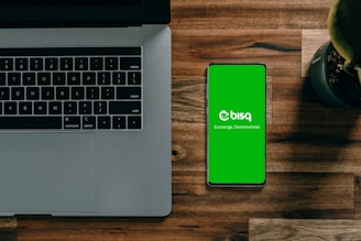 A laptop with a partially visible keyboard situated on a wooden surface next to a smartphone displaying a green screen with the word 'bisq' and the text 'Exchange, Decentralized'. There is also a small potted plant in the top-right corner.