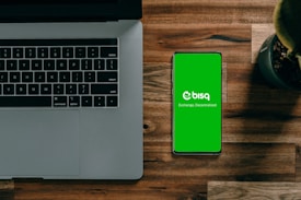 A laptop with a partially visible keyboard situated on a wooden surface next to a smartphone displaying a green screen with the word 'bisq' and the text 'Exchange, Decentralized'. There is also a small potted plant in the top-right corner.