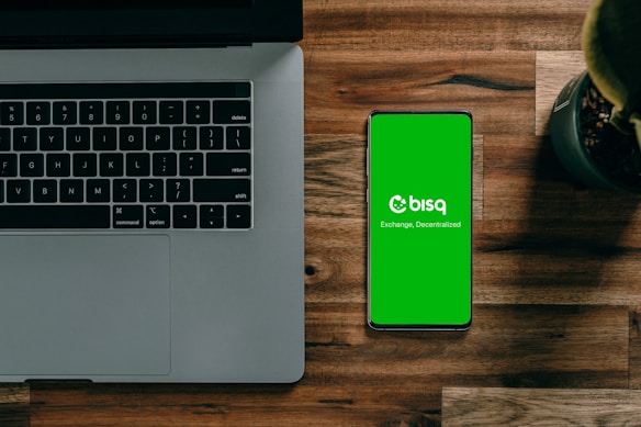 A laptop with a partially visible keyboard situated on a wooden surface next to a smartphone displaying a green screen with the word 'bisq' and the text 'Exchange, Decentralized'. There is also a small potted plant in the top-right corner.