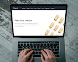 Hands typing on a laptop keyboard with an open webpage displaying information about buying precious metals such as gold, silver, platinum, and palladium. The screen shows an image of gold bars arranged in a pattern. The background consists of a wooden floor with a distressed look.