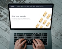 Hands typing on a laptop keyboard with an open webpage displaying information about buying precious metals such as gold, silver, platinum, and palladium. The screen shows an image of gold bars arranged in a pattern. The background consists of a wooden floor with a distressed look.