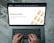 Hands typing on a laptop keyboard with an open webpage displaying information about buying precious metals such as gold, silver, platinum, and palladium. The screen shows an image of gold bars arranged in a pattern. The background consists of a wooden floor with a distressed look.