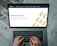 Hands typing on a laptop keyboard with an open webpage displaying information about buying precious metals such as gold, silver, platinum, and palladium. The screen shows an image of gold bars arranged in a pattern. The background consists of a wooden floor with a distressed look.