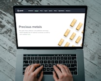 Hands typing on a laptop keyboard with an open webpage displaying information about buying precious metals such as gold, silver, platinum, and palladium. The screen shows an image of gold bars arranged in a pattern. The background consists of a wooden floor with a distressed look.