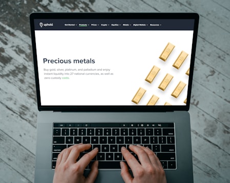 Hands typing on a laptop keyboard with an open webpage displaying information about buying precious metals such as gold, silver, platinum, and palladium. The screen shows an image of gold bars arranged in a pattern. The background consists of a wooden floor with a distressed look.