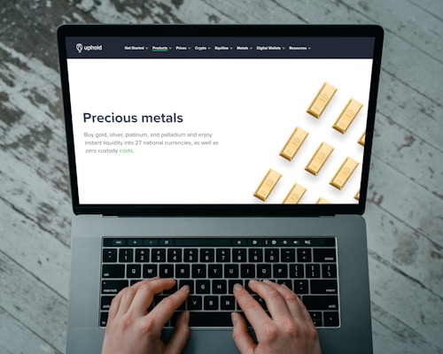 Hands typing on a laptop keyboard with an open webpage displaying information about buying precious metals such as gold, silver, platinum, and palladium. The screen shows an image of gold bars arranged in a pattern. The background consists of a wooden floor with a distressed look.