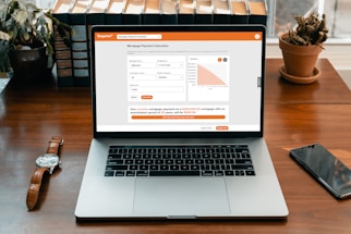 A modern laptop displaying a mortgage payment calculator on its screen. The calculator interface shows input fields for mortgage amount, amortization period, and interest rate, alongside a bar chart. It sits on a wooden desk with a wristwatch, a smartphone, a potted cactus, and a row of books in the background.