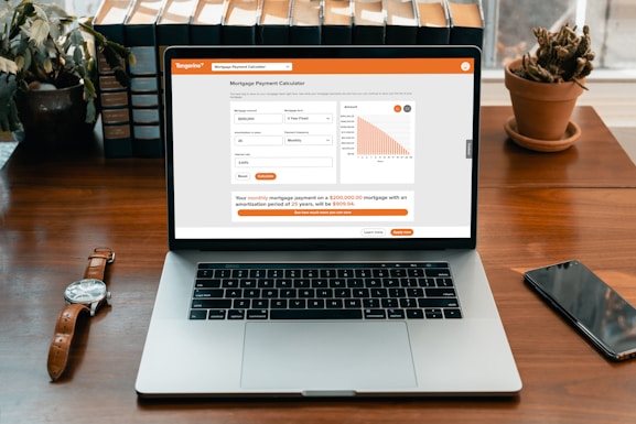 A modern laptop displaying a mortgage payment calculator on its screen. The calculator interface shows input fields for mortgage amount, amortization period, and interest rate, alongside a bar chart. It sits on a wooden desk with a wristwatch, a smartphone, a potted cactus, and a row of books in the background.