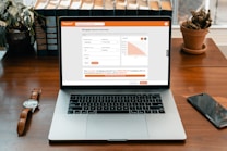 A modern laptop displaying a mortgage payment calculator on its screen. The calculator interface shows input fields for mortgage amount, amortization period, and interest rate, alongside a bar chart. It sits on a wooden desk with a wristwatch, a smartphone, a potted cactus, and a row of books in the background.