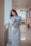 A young woman wearing a long white dress stands in a hallway, holding a blue folder. She leans slightly against a railing, with a gentle and relaxed expression on her face. The corridor is well-lit, with reflections visible on the shiny floor.