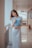A young woman wearing a long white dress stands in a hallway, holding a blue folder. She leans slightly against a railing, with a gentle and relaxed expression on her face. The corridor is well-lit, with reflections visible on the shiny floor.