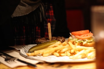 A plate containing a sandwich with tomato slices and lettuce, accompanied by a large serving of French fries and a pickle spear. The food is placed on a piece of paper that resembles newspaper. The background is out of focus, giving the photo a cozy atmosphere.