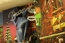 A colorful mural features a stylized mix of architectural elements and a blue humanoid figure with a vintage design. The backdrop includes iconic New York landmarks and neon-like lettering spelling 'Broadway'. Bright colors and artistic renderings make up the background with a patriotic motif including an American flag.