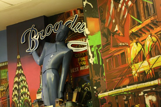 A colorful mural features a stylized mix of architectural elements and a blue humanoid figure with a vintage design. The backdrop includes iconic New York landmarks and neon-like lettering spelling 'Broadway'. Bright colors and artistic renderings make up the background with a patriotic motif including an American flag.
