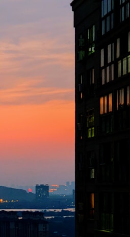 The image captures a cityscape during sunset, with a prominent silhouette of a modern building on the right side. A gradient sky shifts from deep orange at the horizon to soft blue at the top. Distant city buildings and a body of water are partially visible in the background.