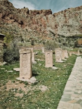 A series of ornate, ancient stone carvings stand in a grassy area against a backdrop of rugged, rocky cliffs. The carvings feature intricate designs and are arranged in a single file. The landscape is natural and historic, with scattered vegetation and a stone pathway on the right.