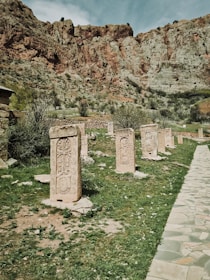 A series of ornate, ancient stone carvings stand in a grassy area against a backdrop of rugged, rocky cliffs. The carvings feature intricate designs and are arranged in a single file. The landscape is natural and historic, with scattered vegetation and a stone pathway on the right.