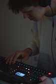 A person wearing headphones is using an electronic music production device. The focus is on their hands interacting with the equipment, surrounded by small, colorful lights on the device.